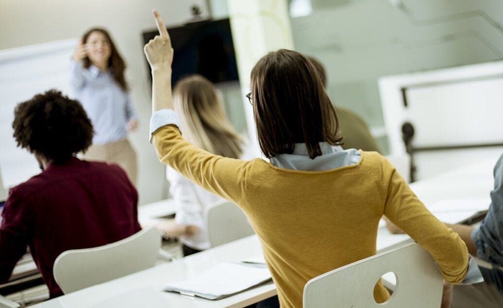 Young adult female student raising her hand while sitting at a long table in classroom, and female teacher pointing at her