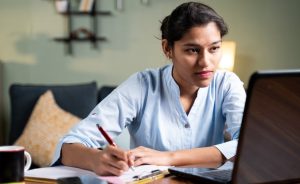 A person is sitting at a table, using a laptop and writing on a notepad.