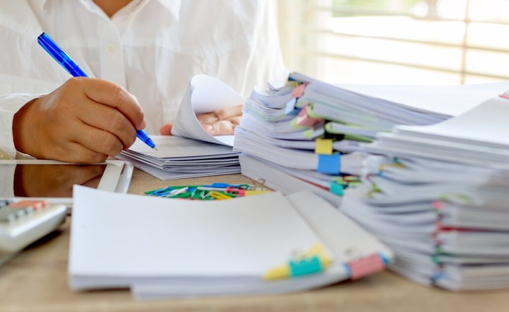 A desk covered in stacks of papers being graded, with paperclips and a teacher writing with a blue pen.