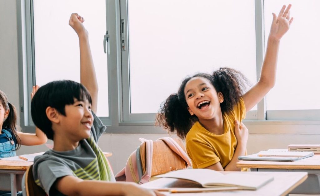 Two smiling elementary students, a boy and a girl, sit at desks with raised hands in a bright classroom.