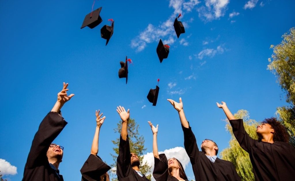 A group of graduates in black gowns throwing their graduation caps in the air against a blue sky.