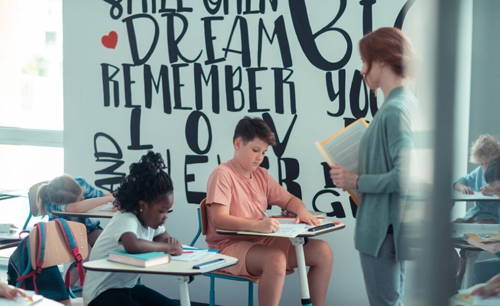Young students working at desks in a classroom with a teacher standing nearby, with inspirational words on the wall.