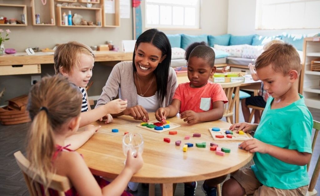 A female preschool teacher helps children with a puzzle activity at a table.
