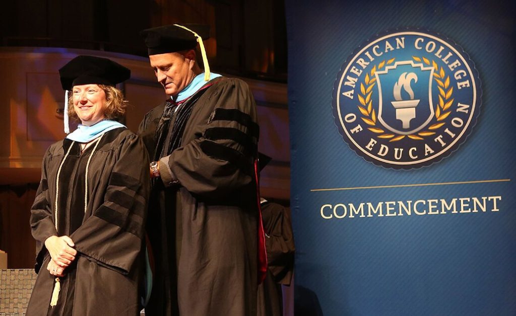 Graduation ceremony with a graduate and faculty member in front of an American College of Education banner