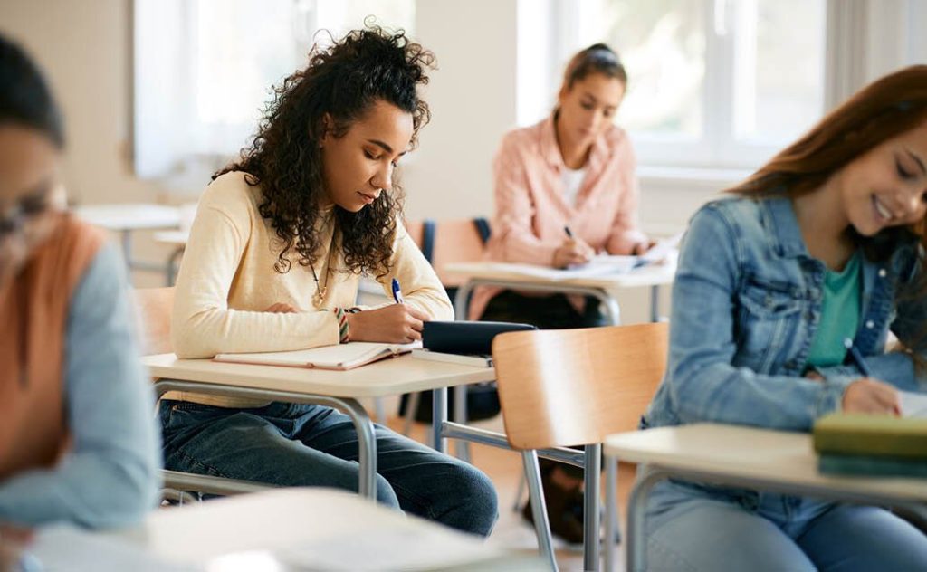 Young female high school student writing in notebook while sitting at desk in classroom