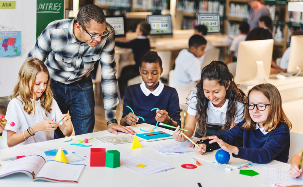 Black male teacher oversees the activities of a diverse group of students, who are sitting at a table working with classroom tools