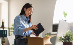 Asian woman packs up her desk, putting a notebook into a brown box