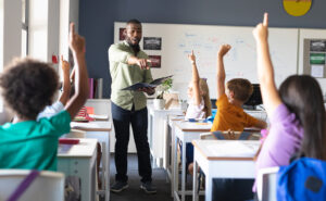 Black male teacher stands in front of several students with their hands raised and calls on one of them