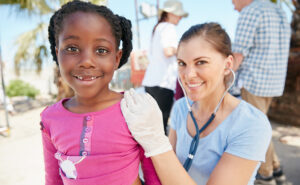 A young Black girl smiles at the camera while sitting on the lap of a white female, who is using a stethoscope to examine the girl at a public health community event