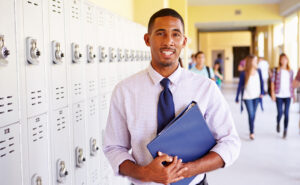 Man in professional dress smiling and holding a binder in front of a row of lockers in a school