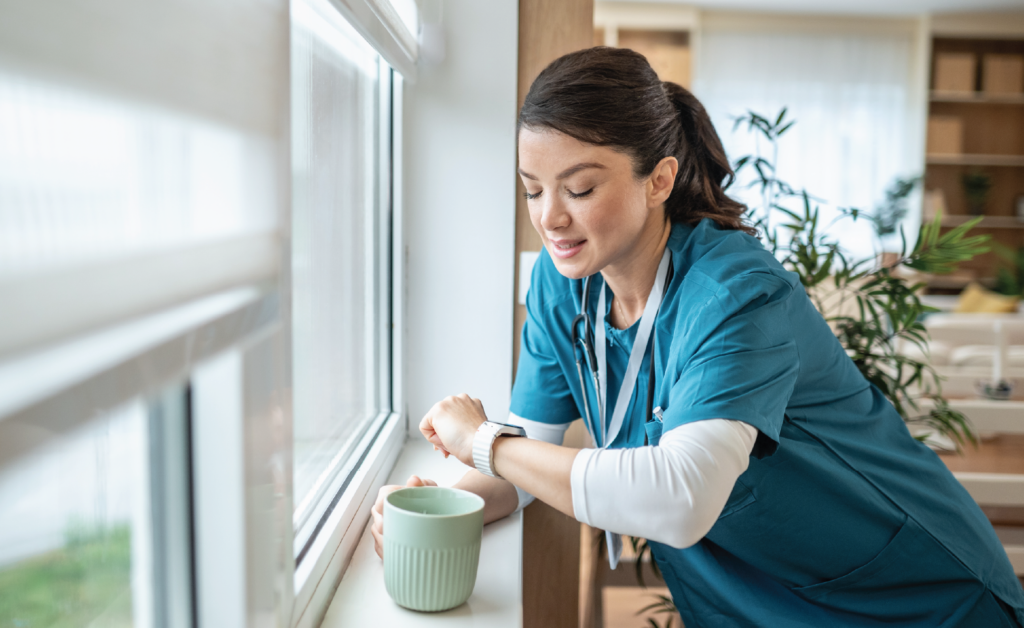 Healthcare professional in scrubs checking her smartwatch while taking a coffee break by a window.
