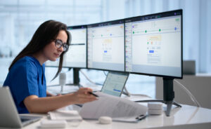 Healthcare worker reviewing patient data across multiple monitors while checking paperwork.
